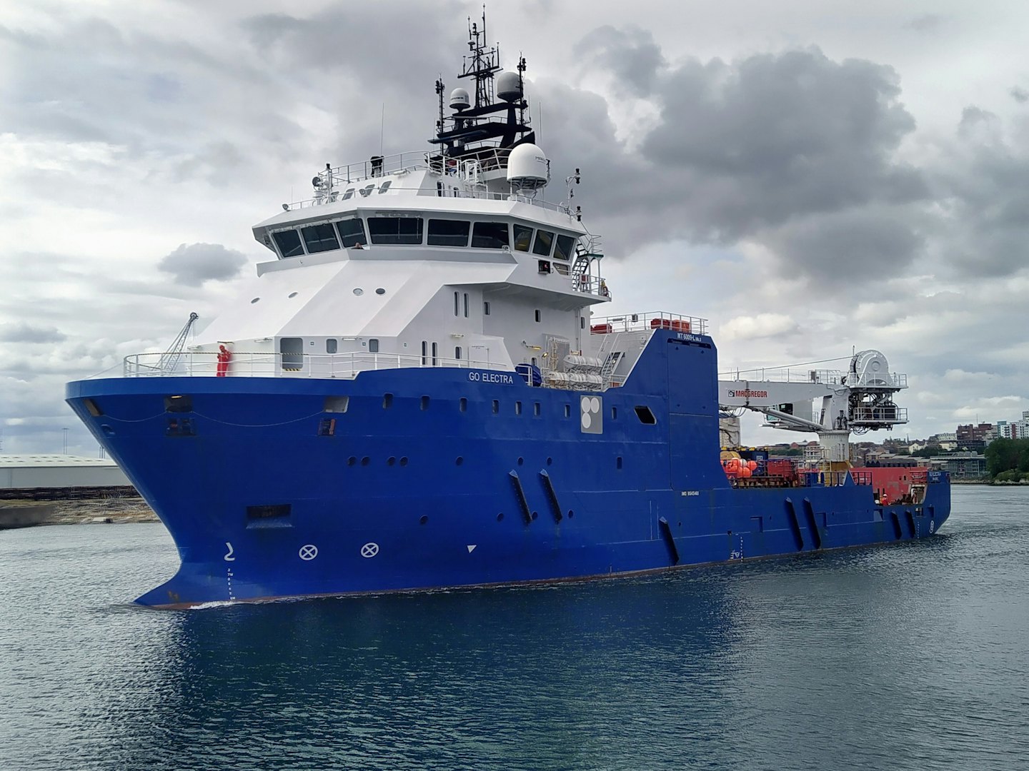 An offshore support vessel travels through a harbor near industrial buildings.